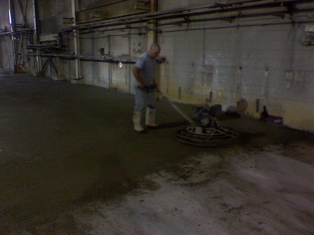 Man using a power trowel on a newly poured concrete floor inside a large industrial building.