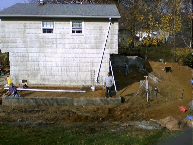 Construction workers near a house with unfinished foundation, surrounded by dirt and equipment.