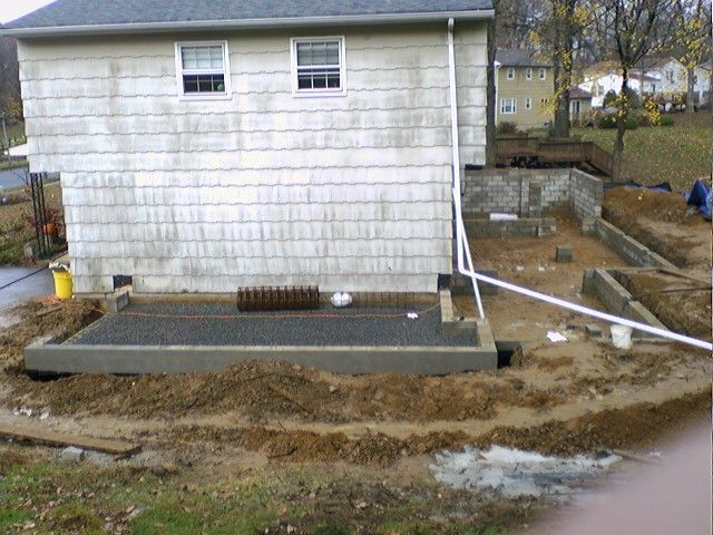Construction site next to a house, concrete patio being built. Brown mud, white walls, cloudy sky.