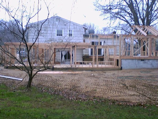 Back view of a house with a wooden frame addition under construction. Dry, brown yard in the foreground.