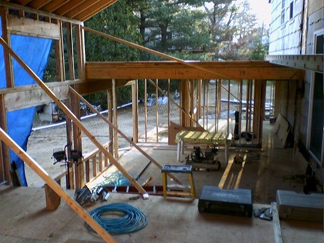 Construction site: wooden framing of a deck or porch addition. Tools and materials are scattered. Sunny outdoor setting.