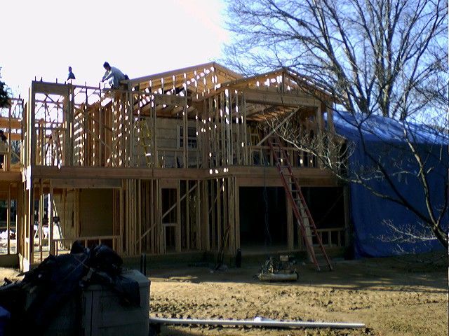 Construction of a two-story house with wooden framing. A worker is on the roof.