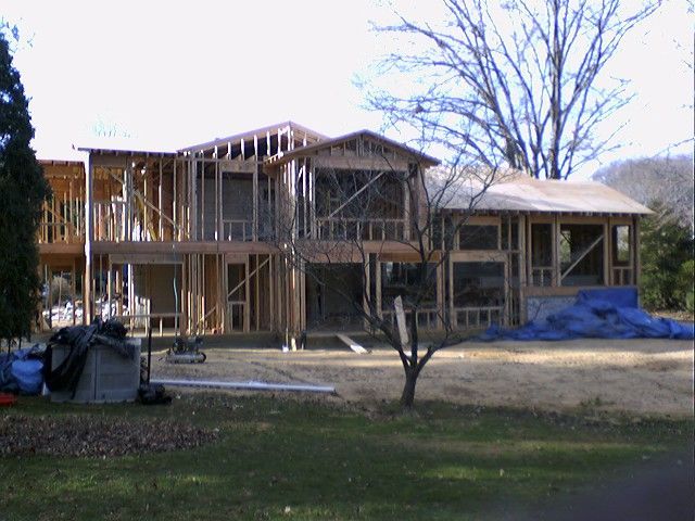 House under construction; wooden frame, windows, on grassy lot, tarp covered materials, bare trees.