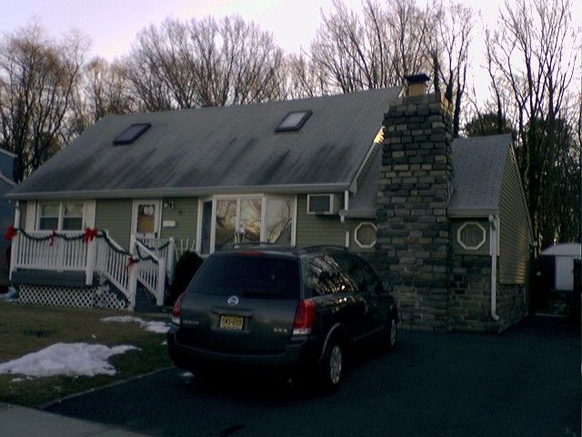Green house with SUV parked in front, stone chimney, snow on lawn.