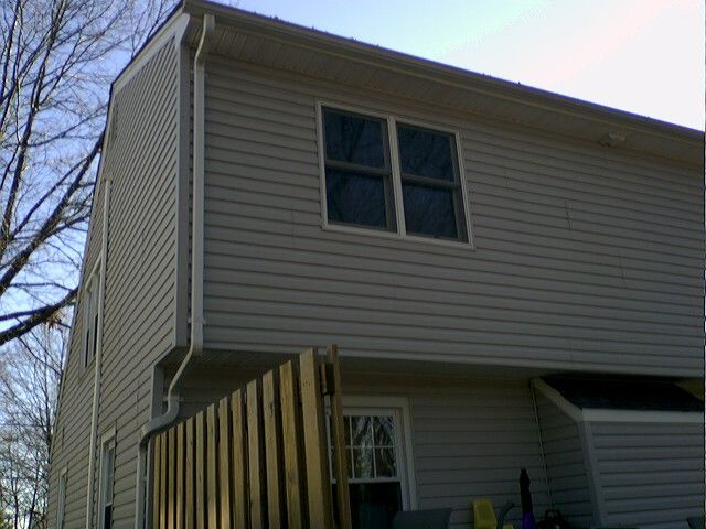Two-story beige siding house with windows, gutter, and part of a wooden fence on a sunny day.