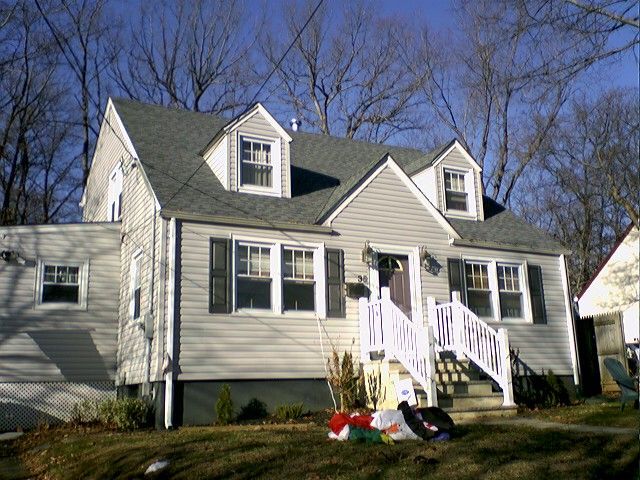 Two-story gray house with white trim, black shutters, three dormers, and a white porch with a blue sky background.