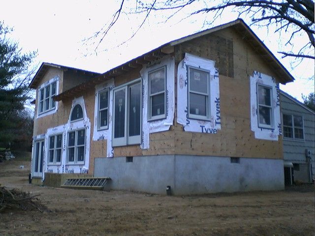 House under construction with plywood siding, windows framed with white paper, and a concrete foundation.