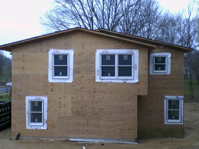 House under construction with plywood siding, white window frames, and cloudy sky.