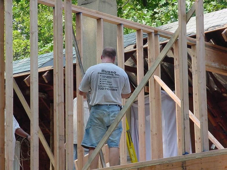 Man in a t-shirt working on wooden framing; chimney in background, trees visible.