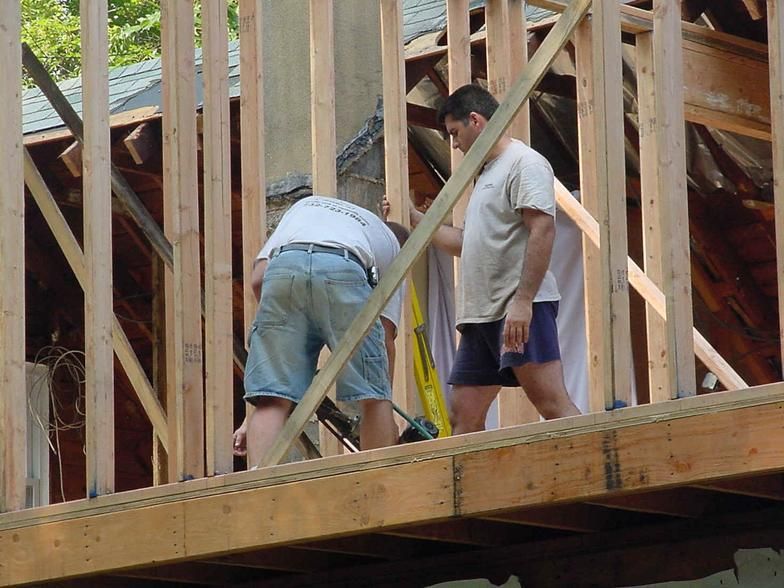 Two men building a wooden structure. One measures, the other assists, both are on a roof. Sunny day.