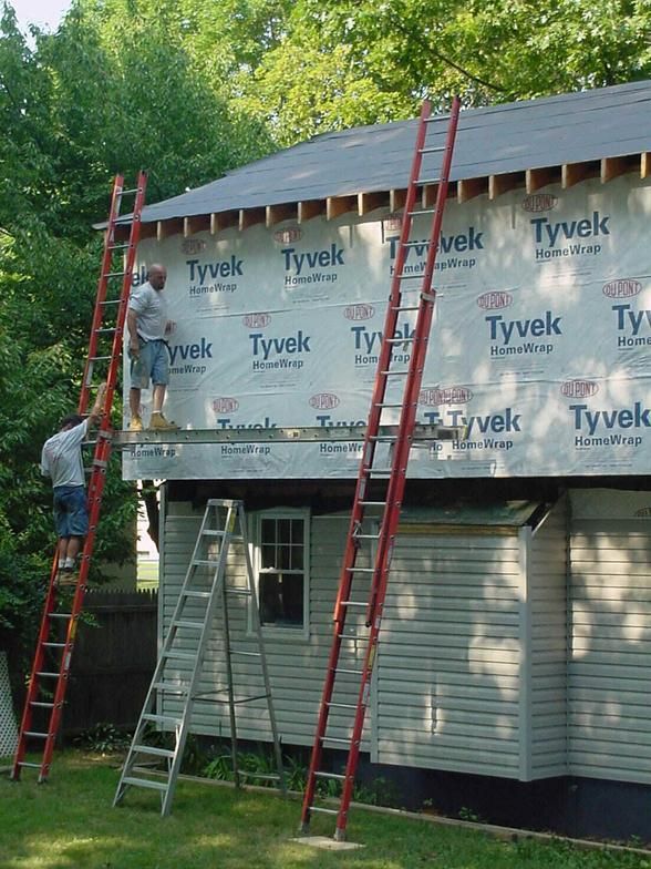 Workers on ladders applying Tyvek to a building's exterior; green grass and trees in the background.