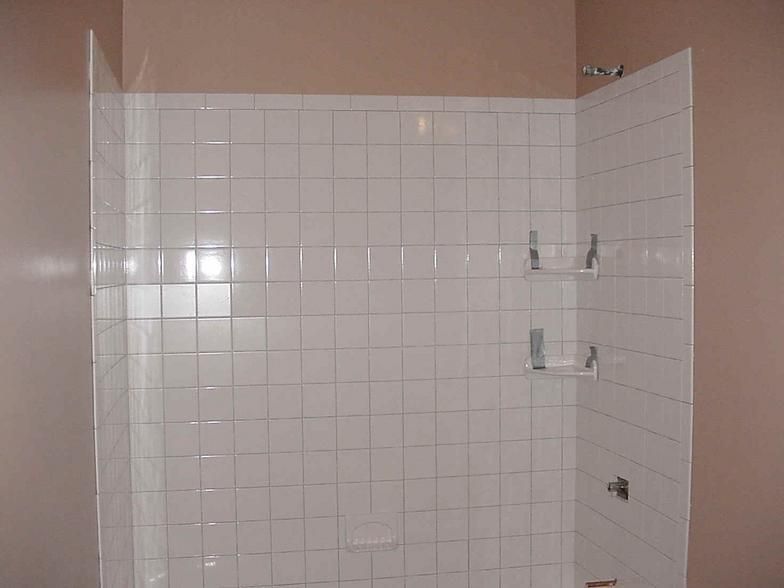 White-tiled shower enclosure with a showerhead and two built-in shelves against a light brown wall.