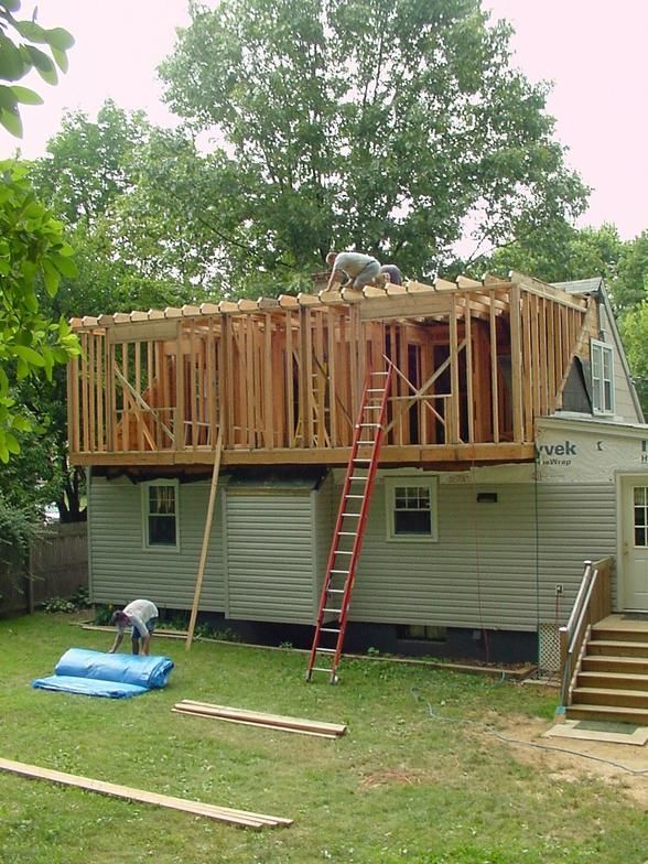 Construction of a second story addition on a house. Workers on the frame. Green grass, blue tarp.