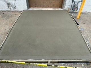 Freshly poured concrete ramp outside a doorway, bordered by wooden forms and yellow caution tape.