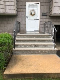 Three concrete steps lead to a white door with a wreath. Black iron handrails flank the steps.