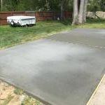 A newly poured concrete pad in a backyard. A trailer is parked in the background, and grass surrounds the pad.