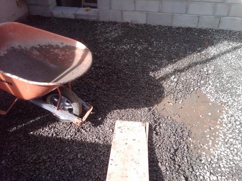 Wheelbarrow with gravel next to concrete block wall; wet gravel and small puddle on ground.