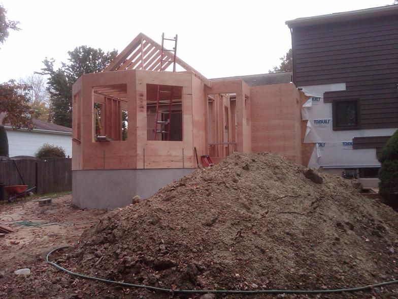 Construction of a home addition; unfinished wooden walls, roof rafters, dirt pile, and house with brown siding.