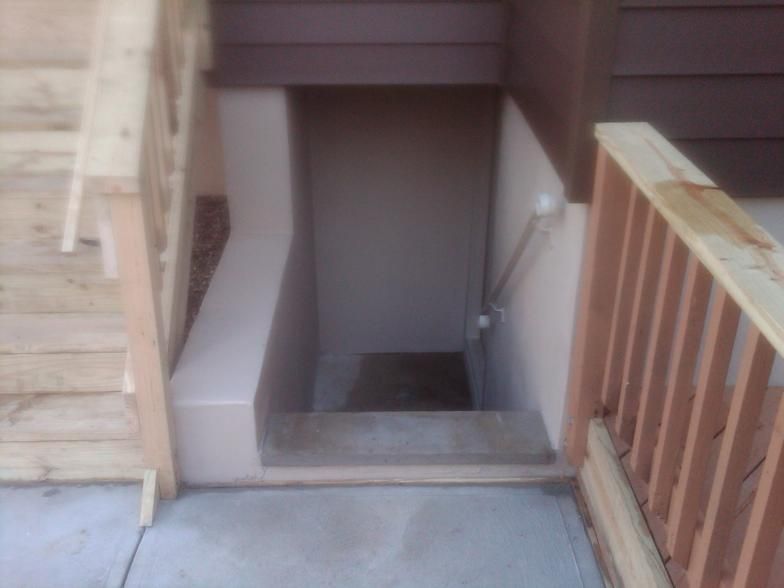 Entryway with stairs and handrails leading down to a basement door. Concrete and tan/brown siding.