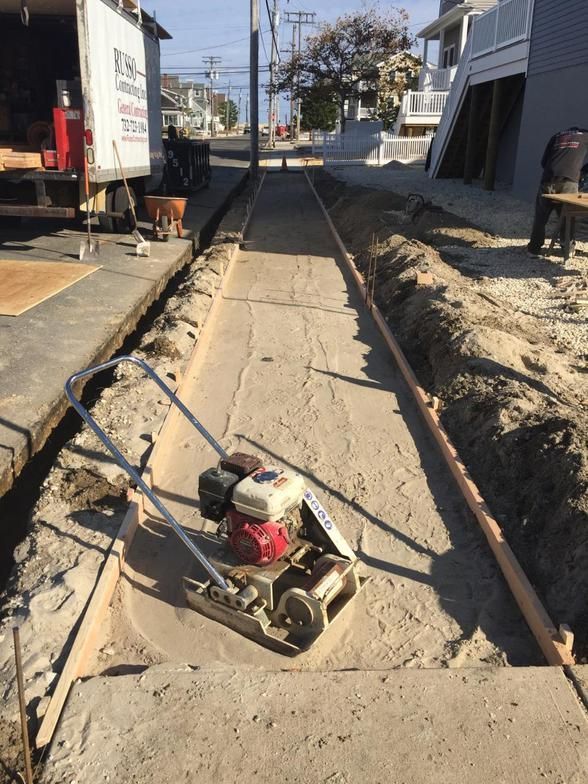 A vibratory plate compactor compacting sand in a sidewalk trench, street in the background.