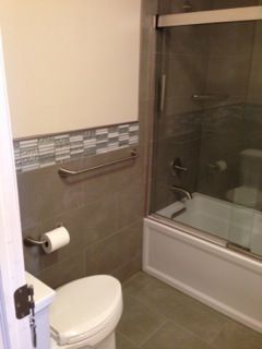 Bathroom with toilet, bathtub, and glass shower door. Gray and white tiles and walls.
