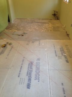 Room with flooring work in progress; cement floor visible, some overlaid with Durock sheets, green walls.