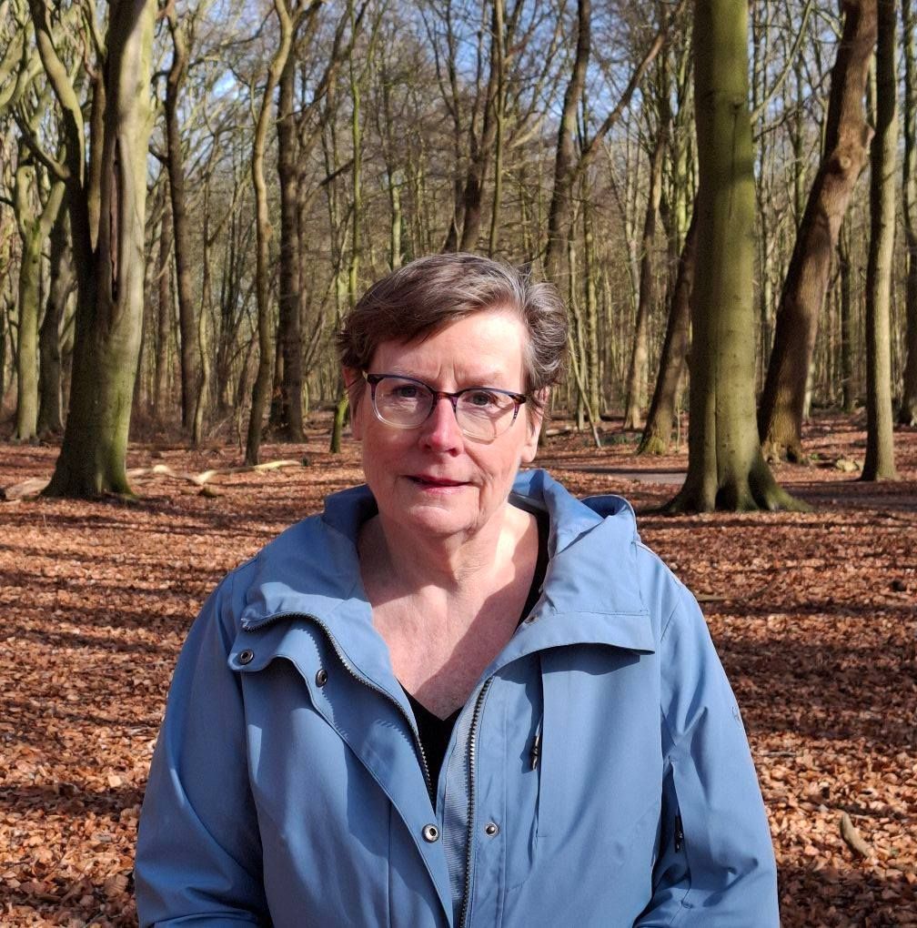 Een vrouw met een bril en een blauw jasje staat in het bos