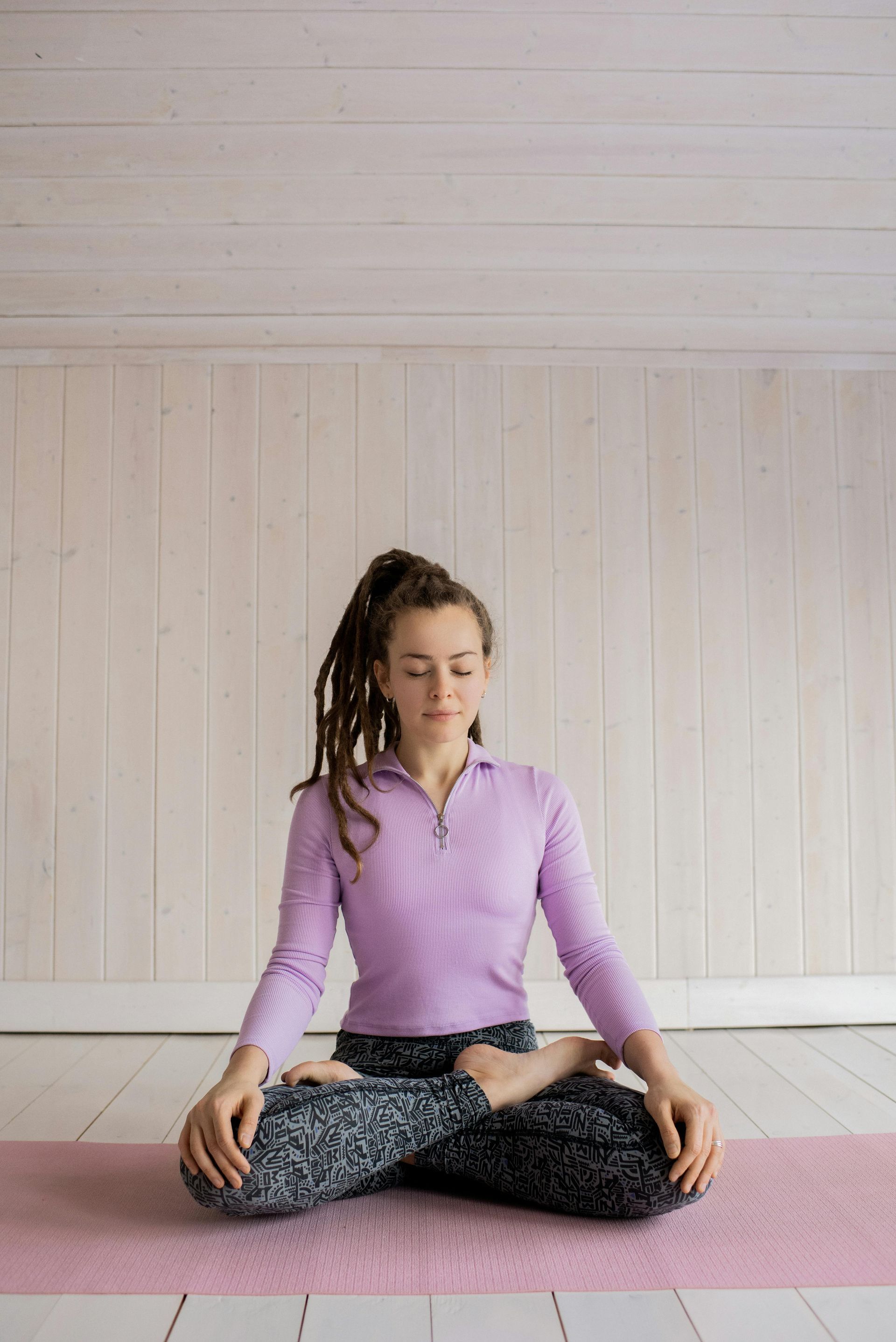 Mujer en posición de loto, con los ojos cerrados, sobre una esterilla rosa en una habitación de paredes blancas, practicando yoga.