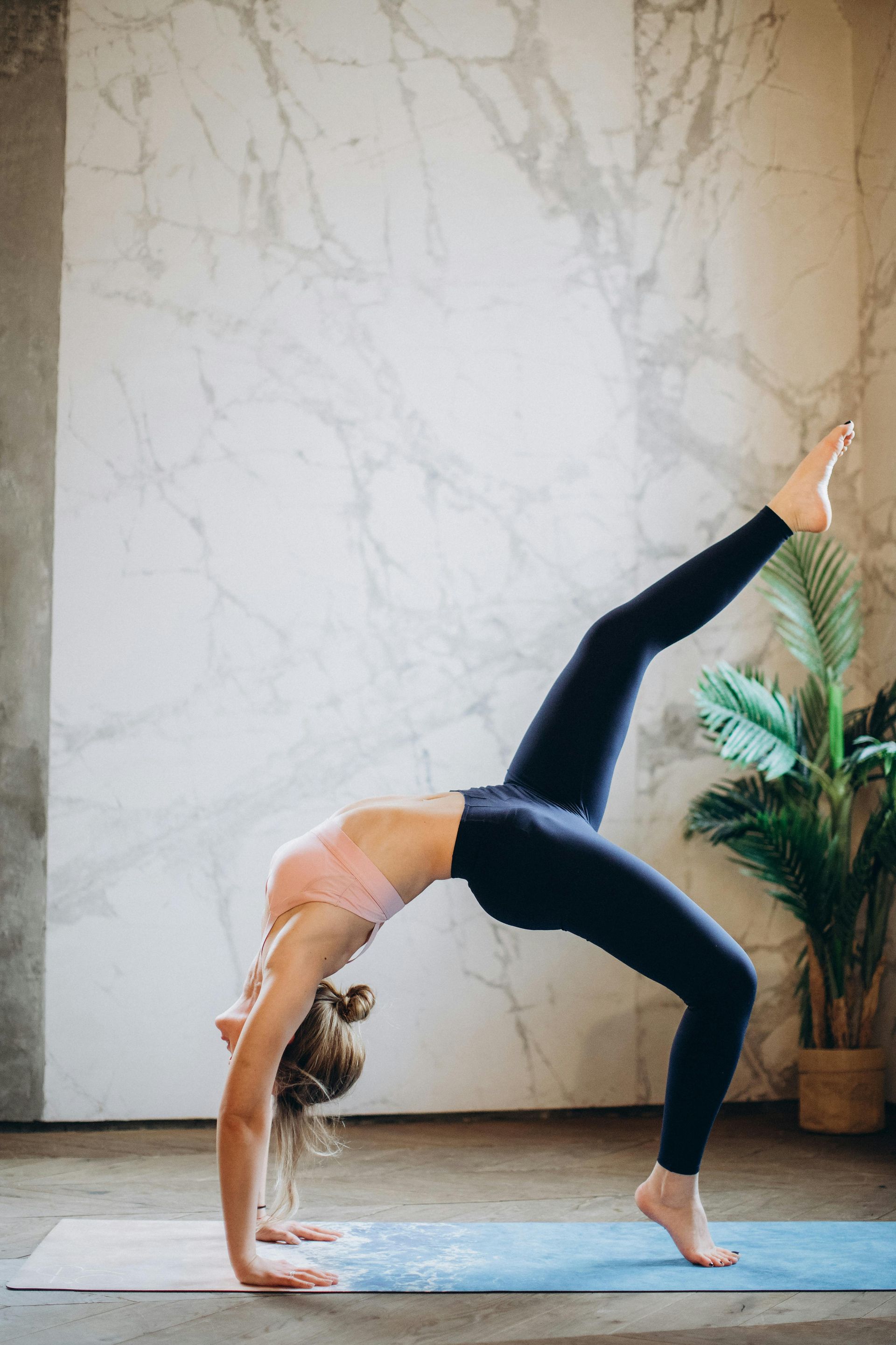 Mujer en pose de yoga, espalda arqueada, una pierna levantada, sobre una colchoneta en el interior con pared de mármol y planta.