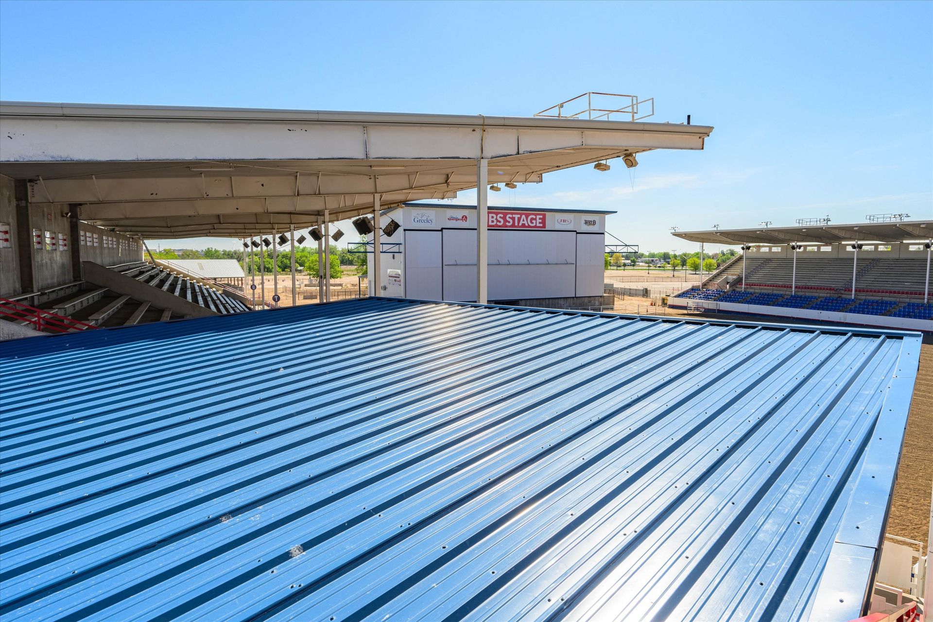 Blue metal roofing with bleachers and a concession stand under a stadium's roof on a sunny day.