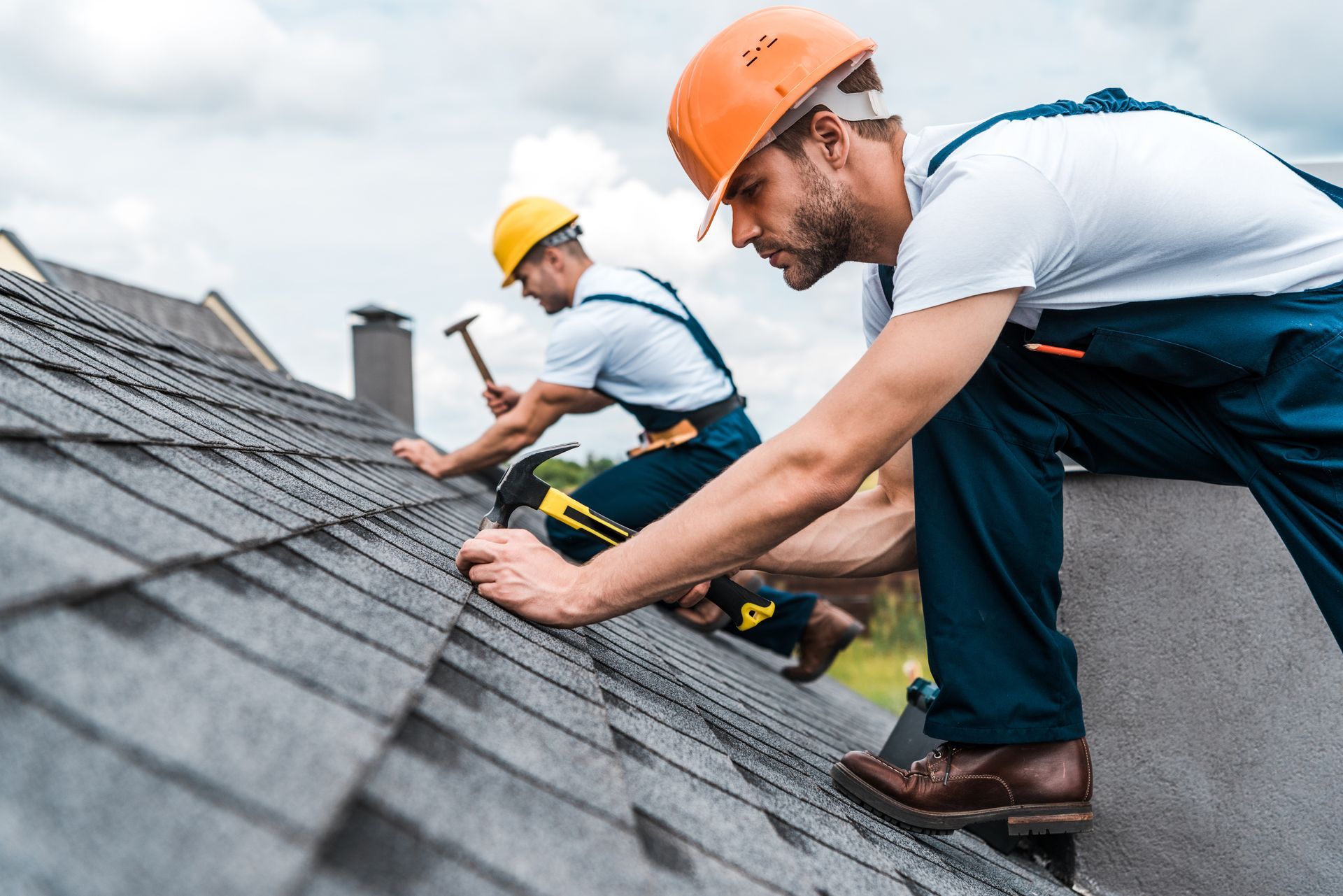 Two roofers wearing helmets and overalls are performing a roof repair service on a house.