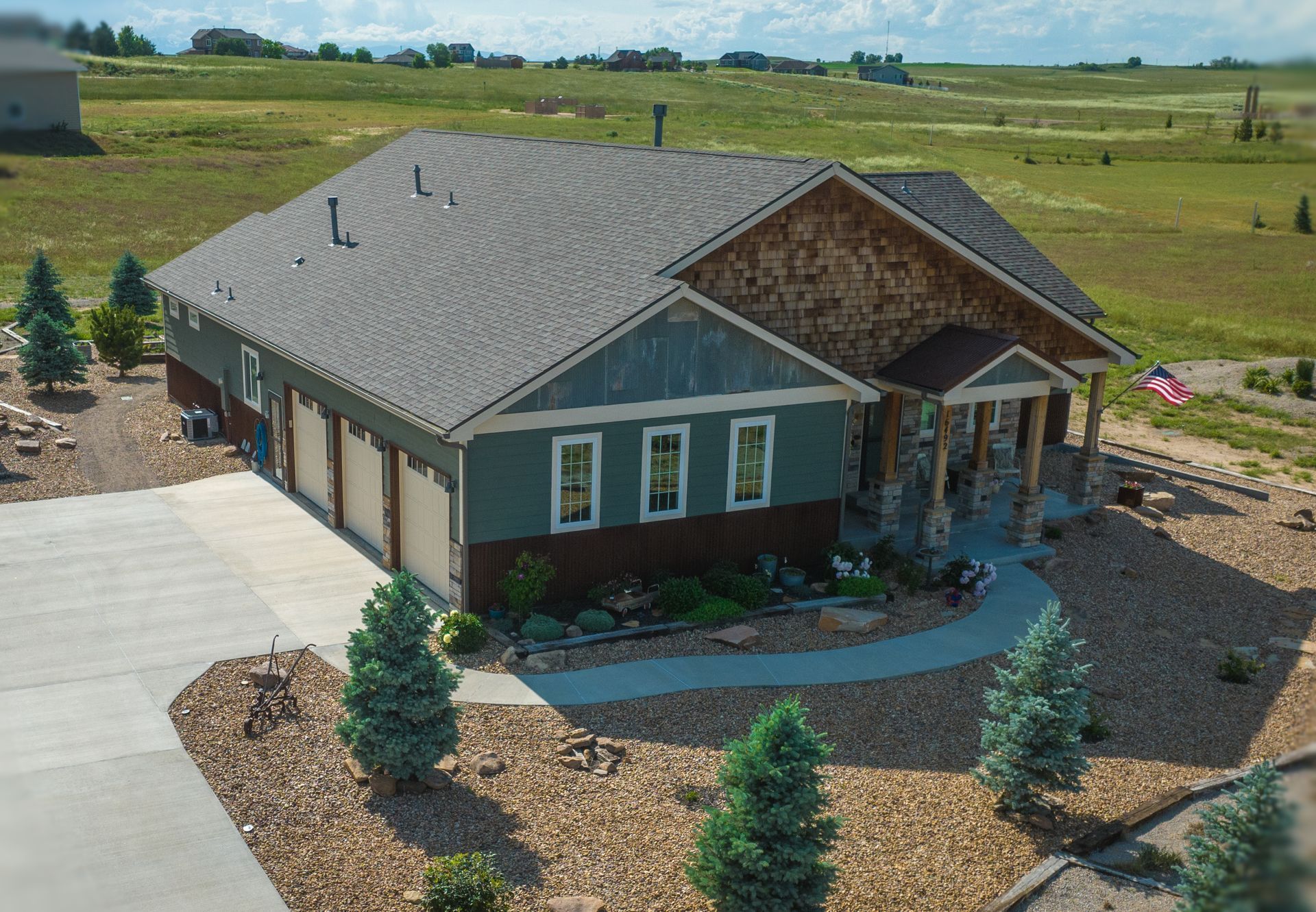 House with a gray roof, blue siding, brown trim and garage doors. Located in a rocky area with a field.