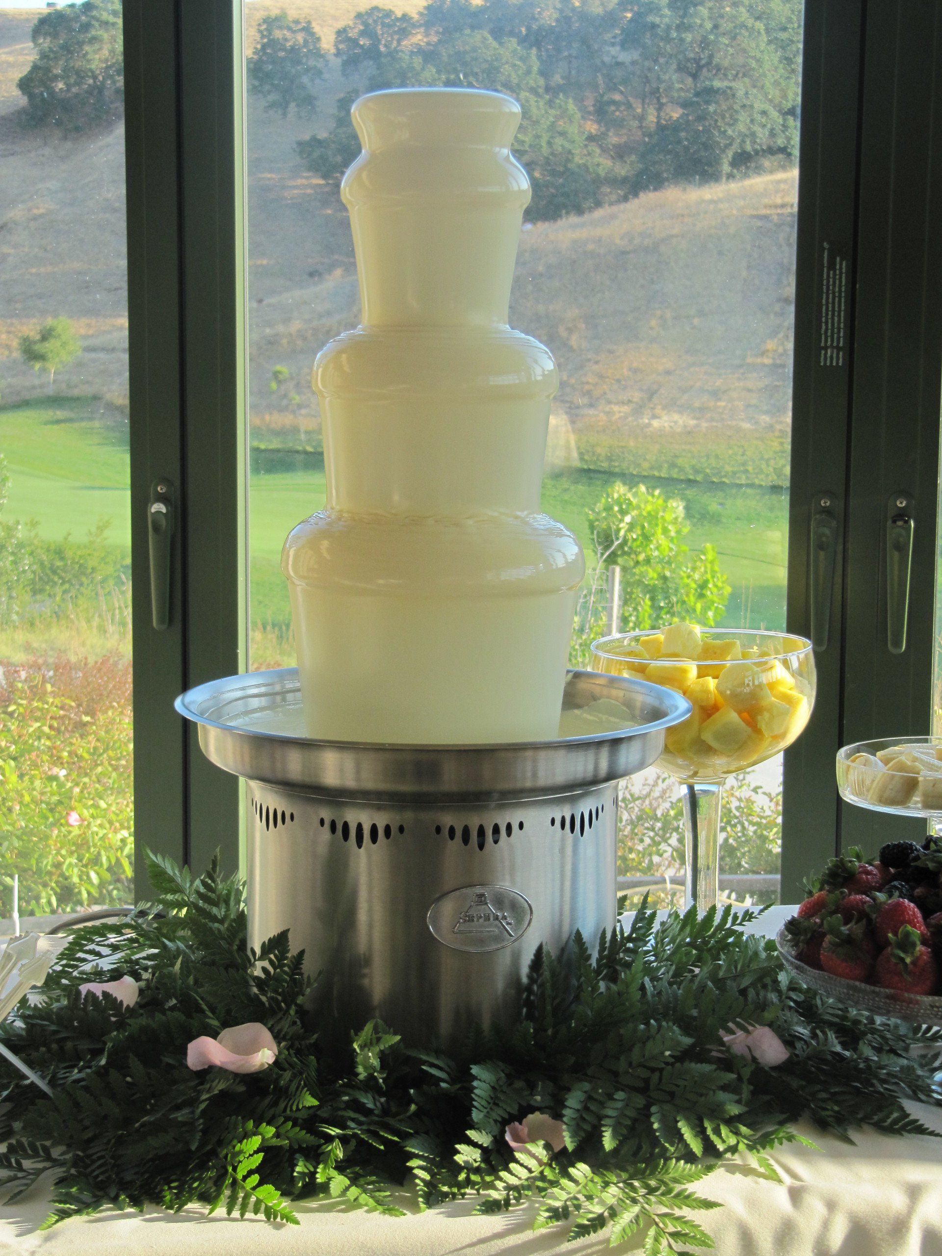 A white chocolate fountain is sitting on a table in front of a window.
