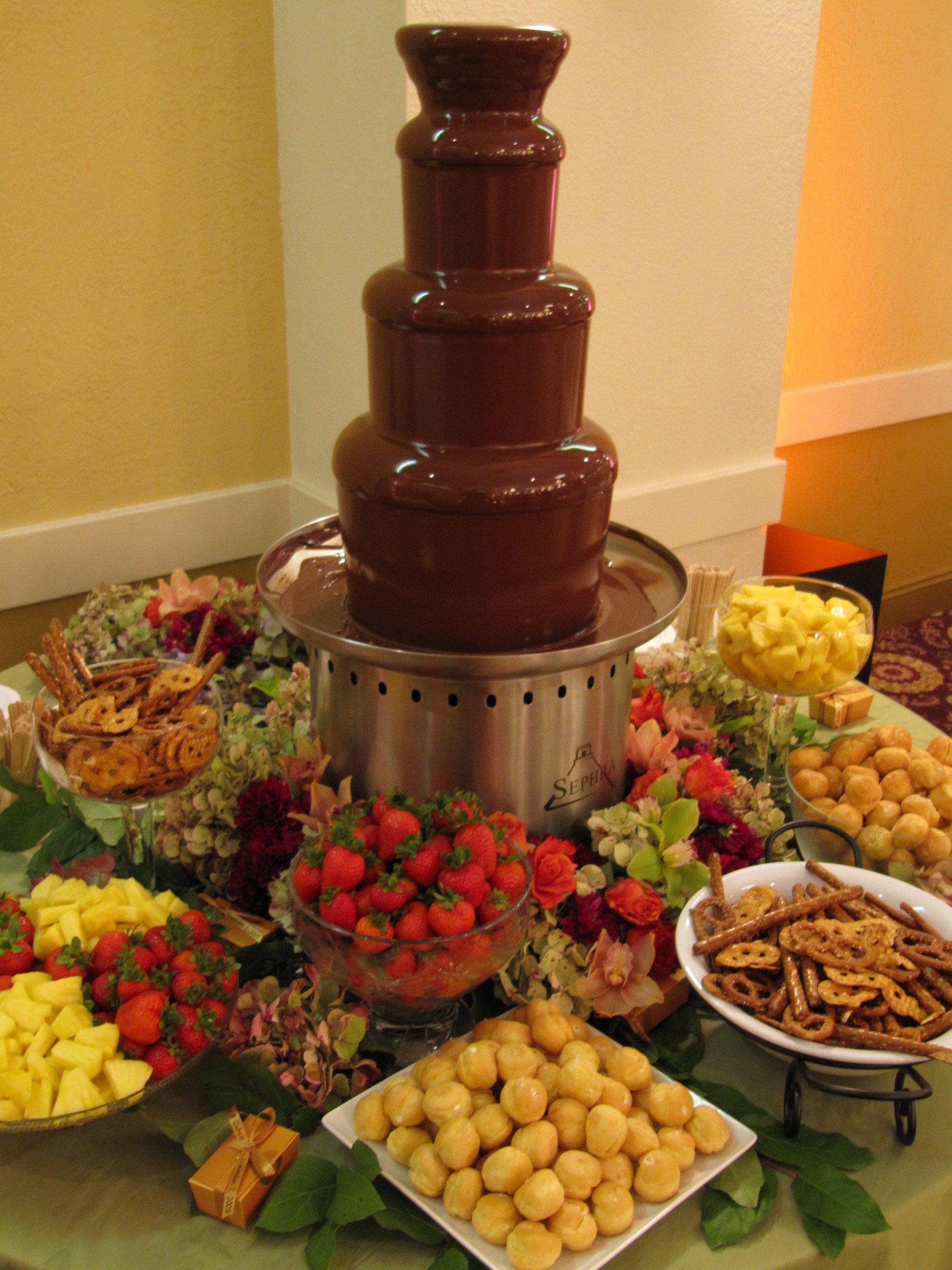 A chocolate fountain is surrounded by fruit and pretzels on a table