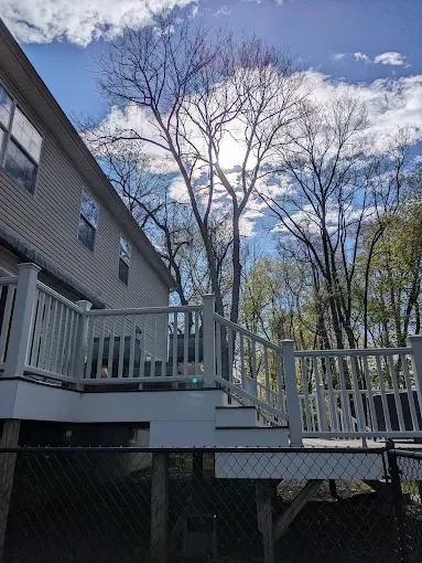 A large white deck with stairs leading up to it is in front of a house with trees in the background.