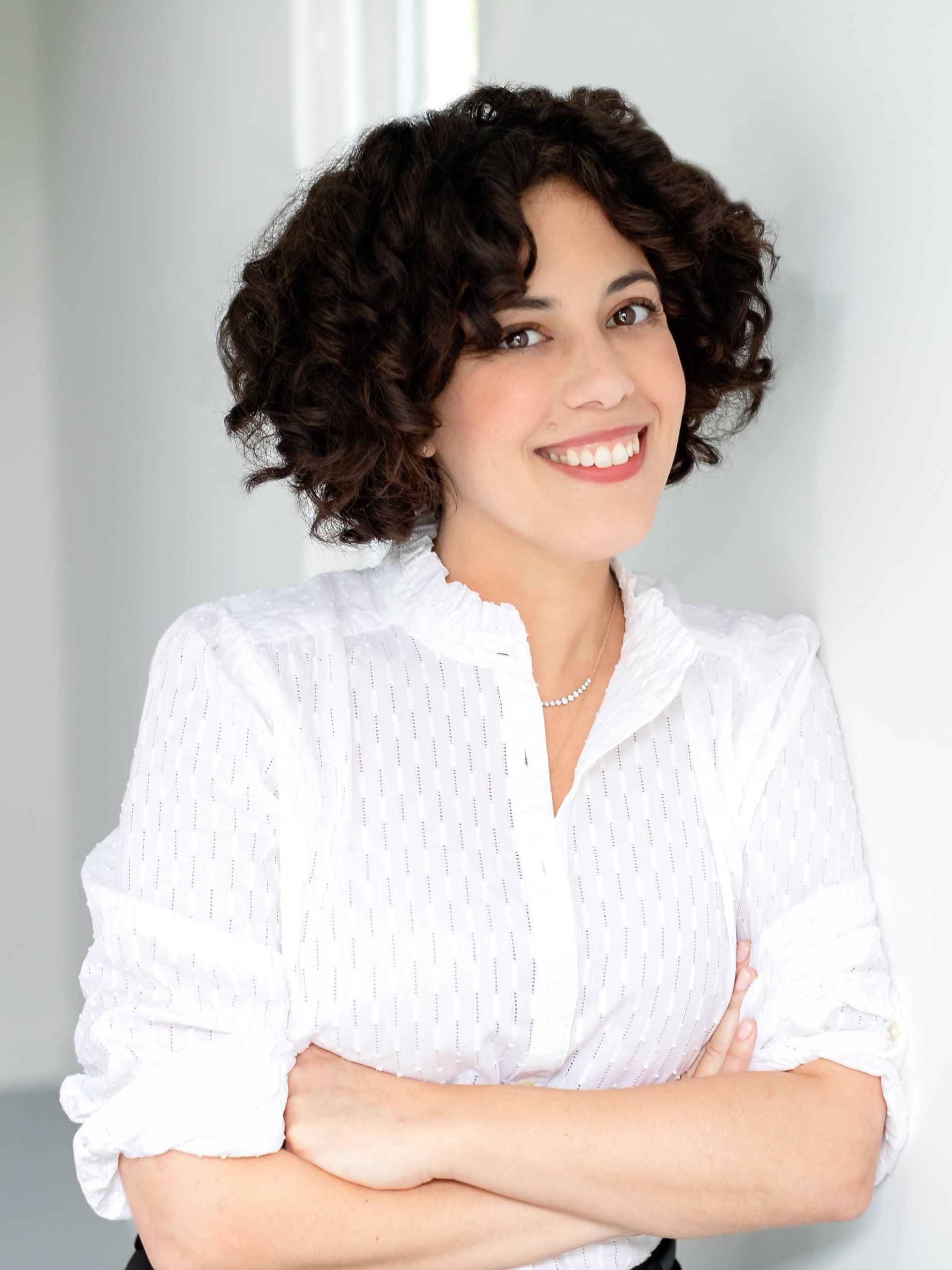 A woman with curly hair is wearing a white shirt and smiling with her arms crossed.