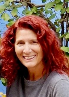 A woman with red hair is smiling in front of a tree.