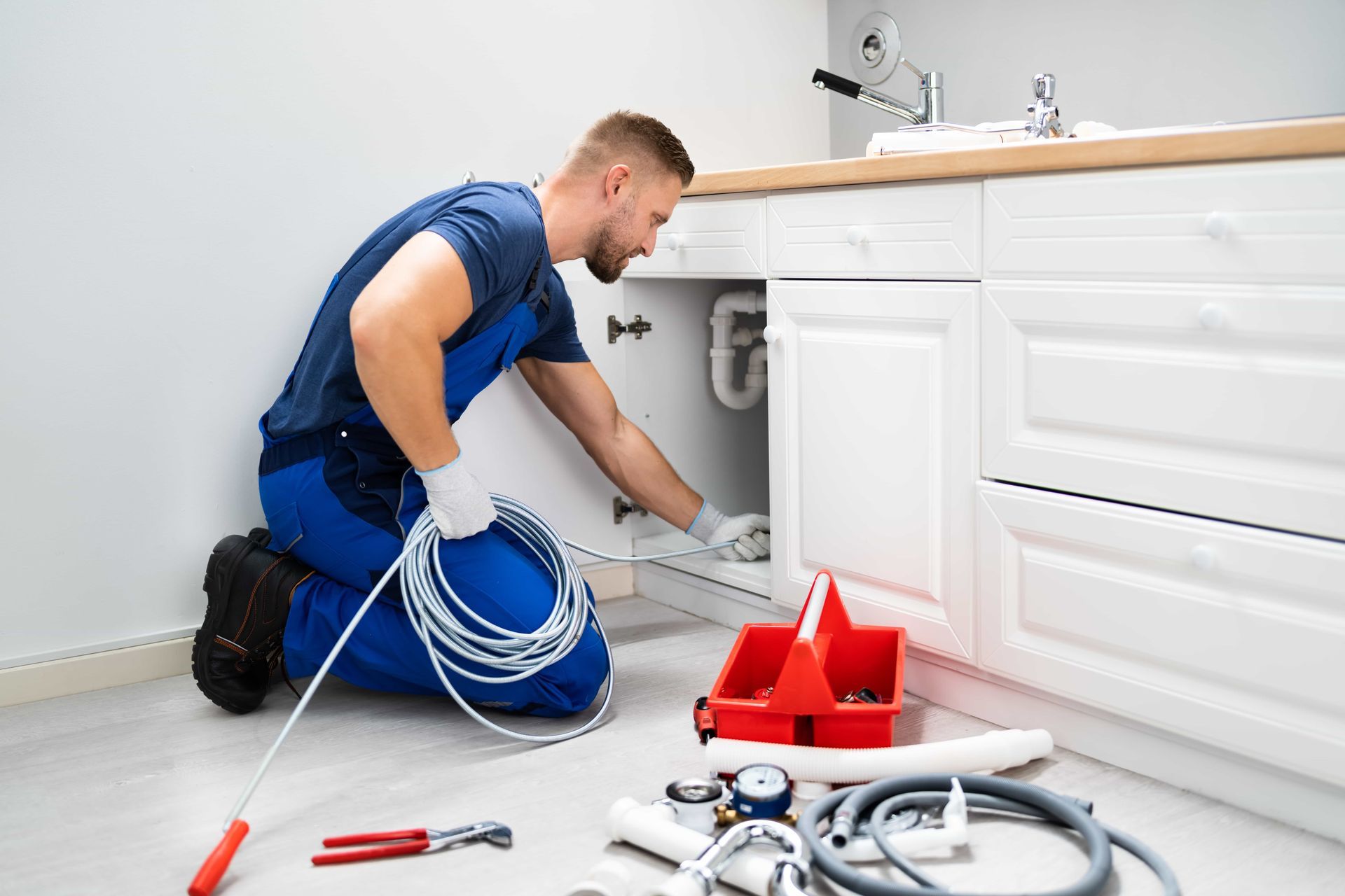 Plumber kneeling under a kitchen sink, using a snake. White cabinets, red toolbox, and tools visible.