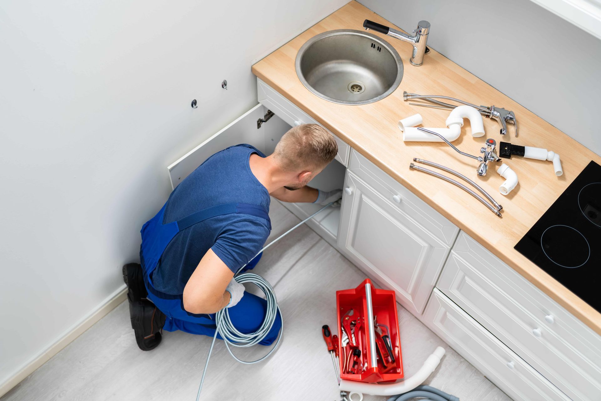 Plumber working under a kitchen sink, using a snake tool; tools and parts on counter nearby.