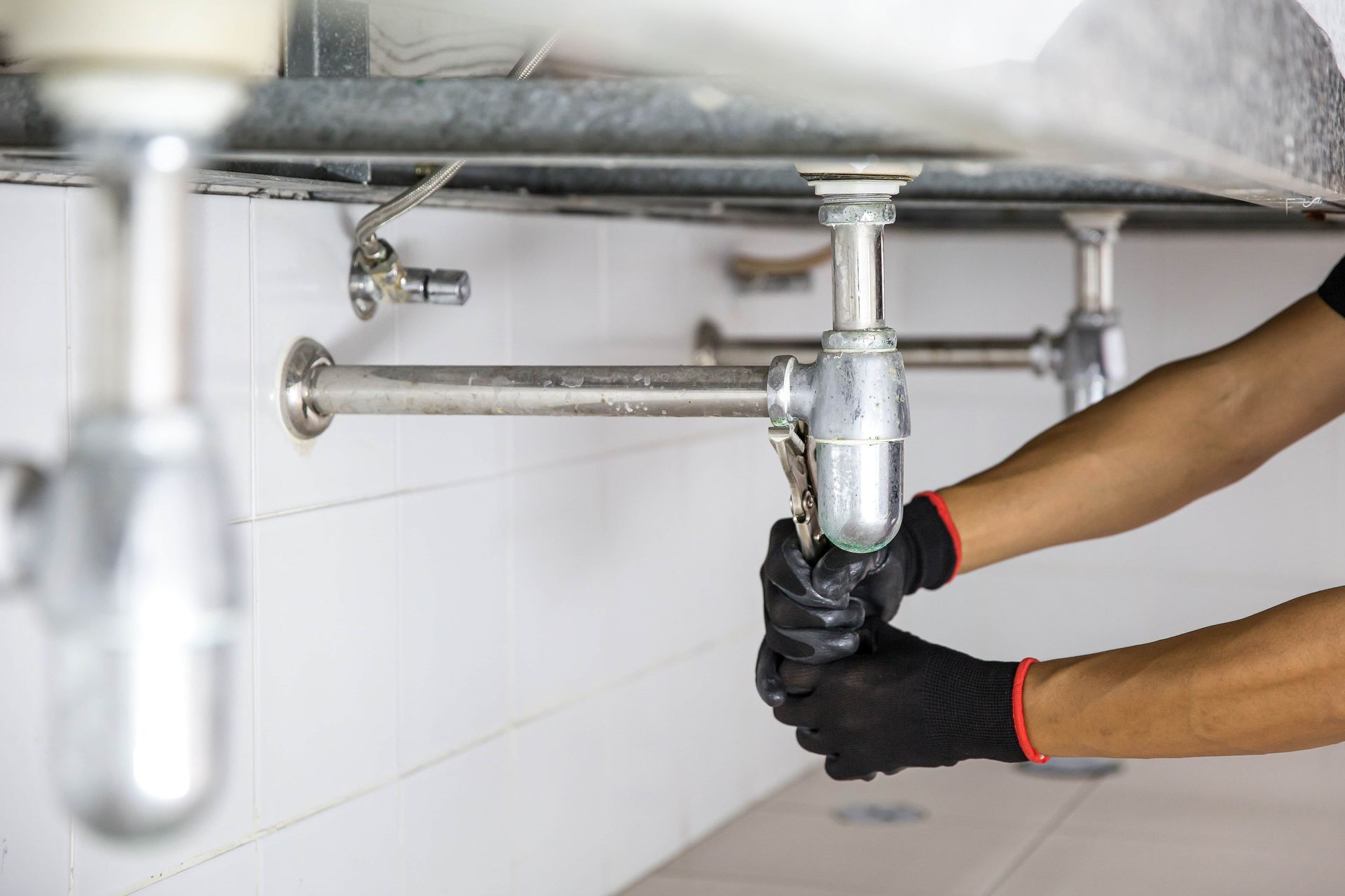 Hands wearing black gloves use a wrench to work on plumbing under a sink.