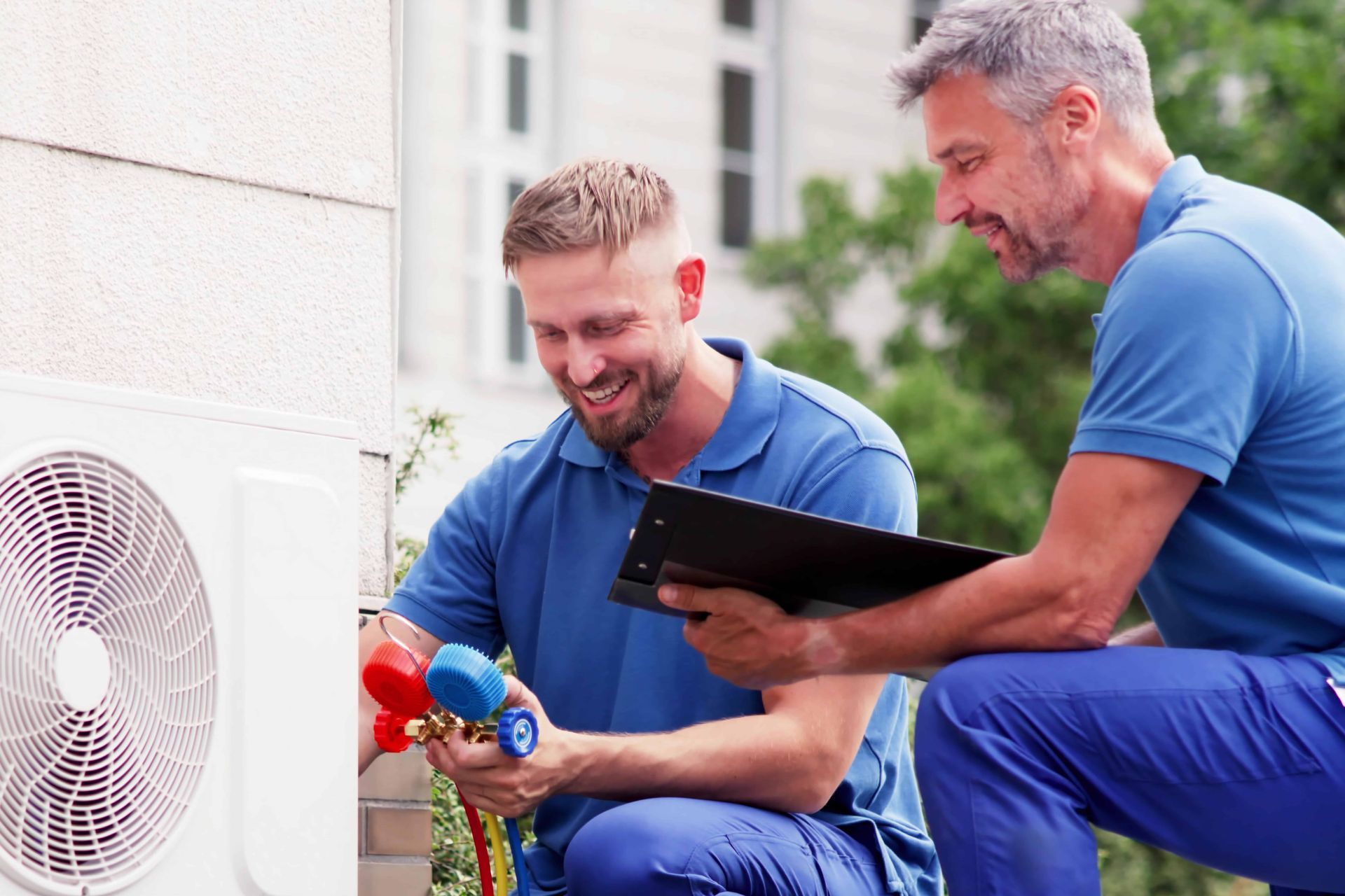 Two technicians in blue shirts inspect an AC unit, one with gauges, one with a clipboard, smiling.