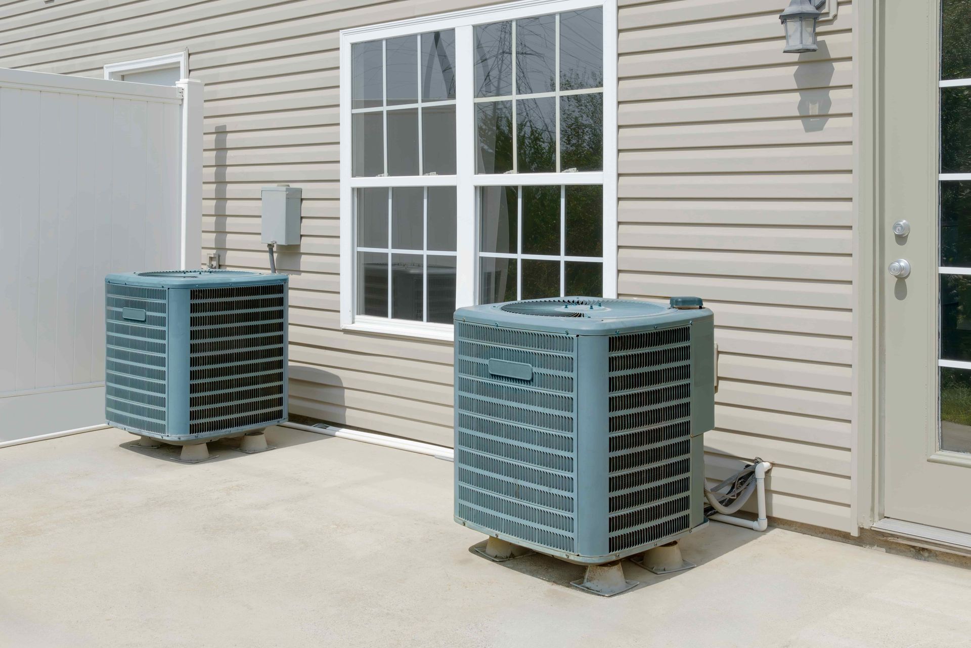 Two air conditioning units on a concrete patio next to a house with a window and door.