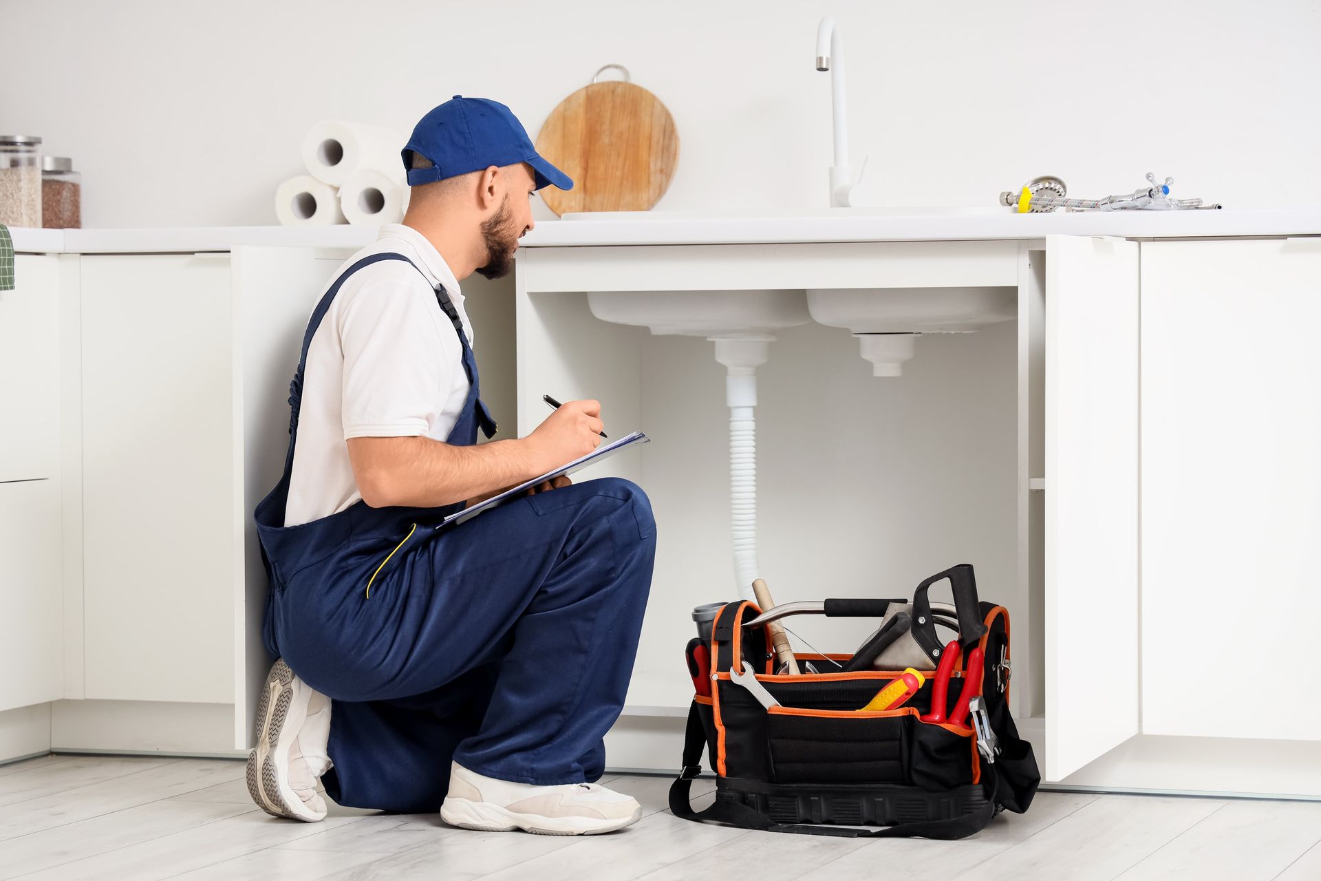 Plumber kneeling under a kitchen sink, writing on clipboard, with a toolbox nearby.