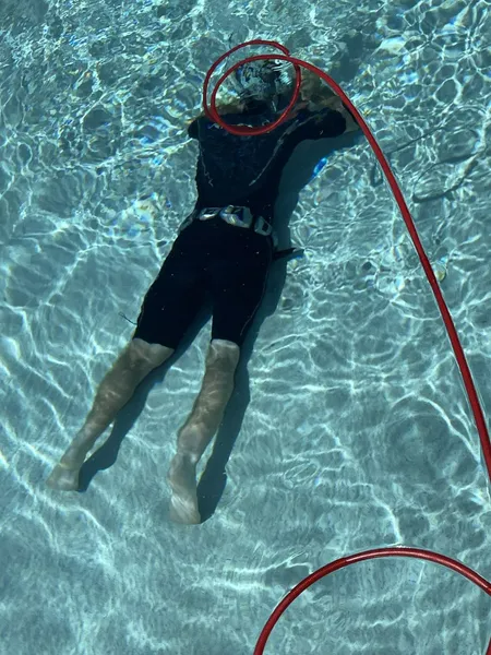 Person in black swimsuit underwater near red tubing in pool.