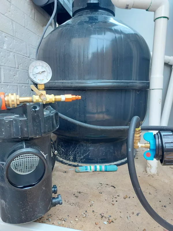 Pool filter system: black pump, pressure gauge, sand filter, and hoses against a brick wall.