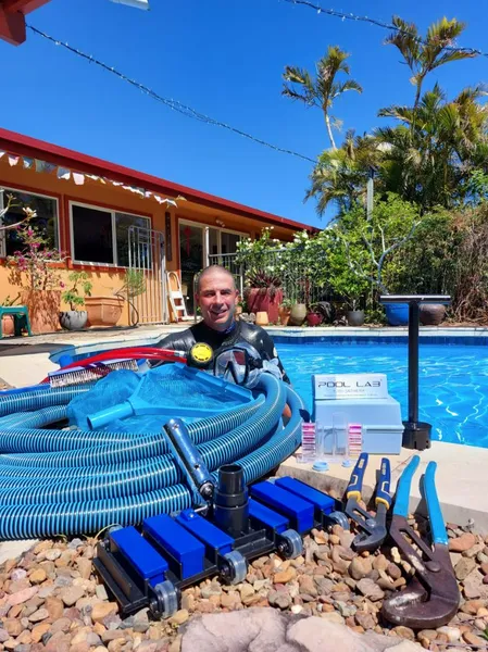 Man in wetsuit by pool with pool cleaning equipment. Sunny outdoor setting.