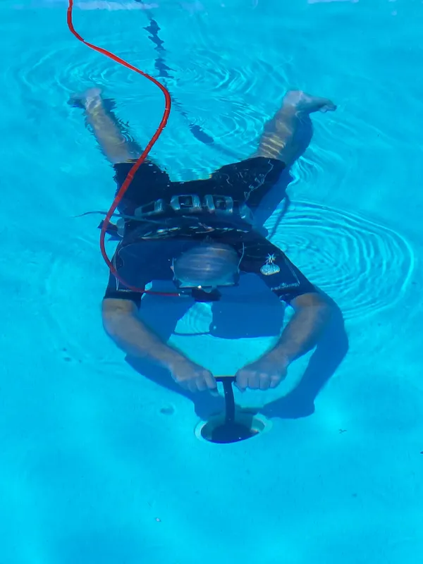 Person underwater in pool using a pool cleaner. He wears a diving mask and holds the cleaner's handle.