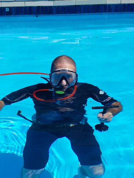 Man in scuba gear underwater in a pool, facing the camera. Blue water, black wetsuit, and mask.