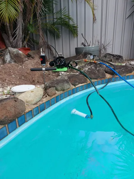 Pool vacuum in operation, blue water, green and black equipment near pool edge, gray fence background.