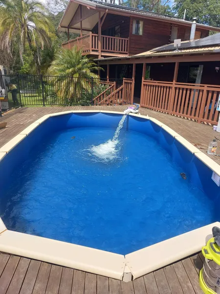 Blue pool with water fountain, surrounded by a wooden deck and brown house.
